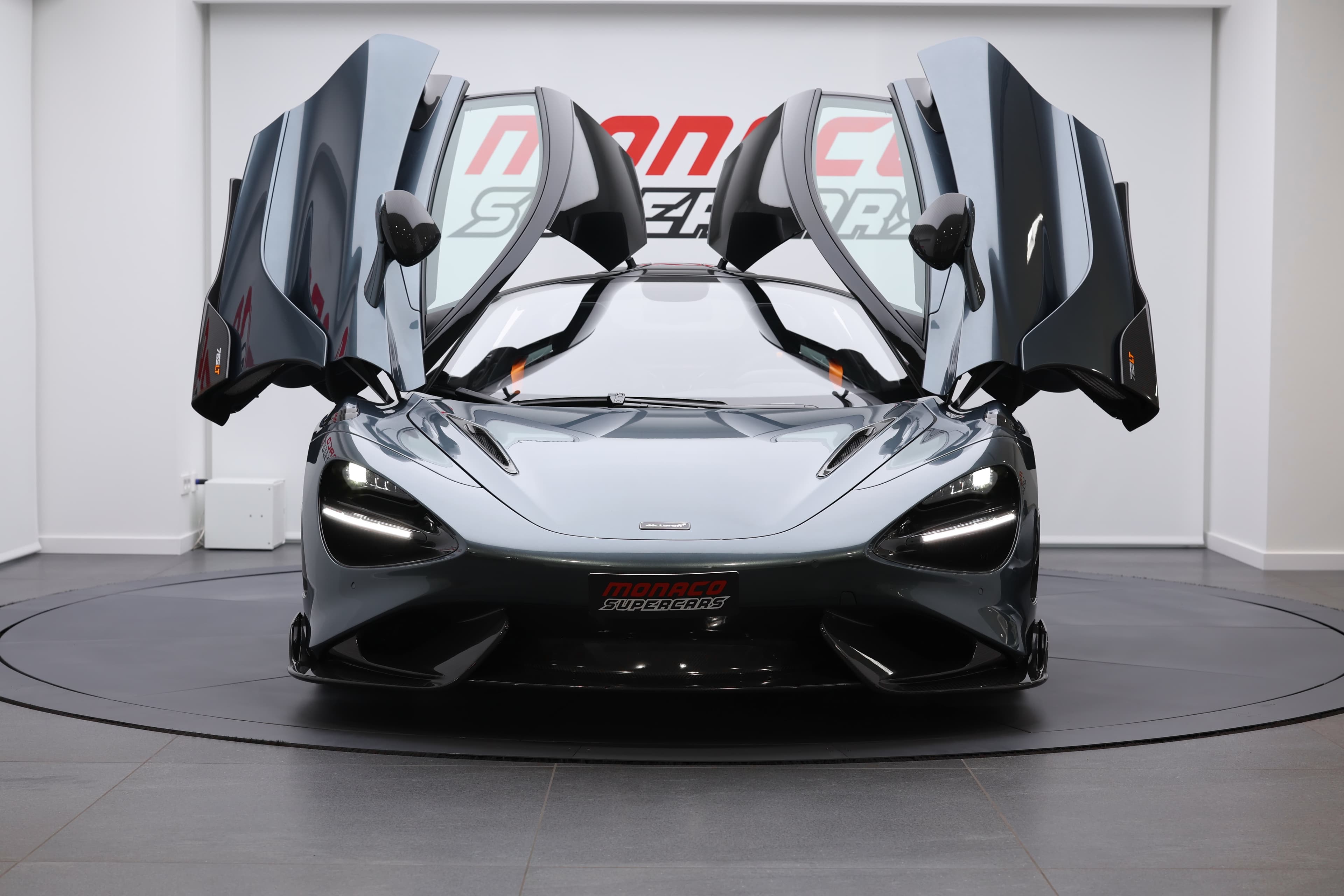 L'image montre la face avant d'une McLaren 765LT avec les portes papillon ouvertes, mettant en avant le design saisissant de la voiture et l'architecture unique de son habitacle, tout en soulignant son caractère sportif et élégant.