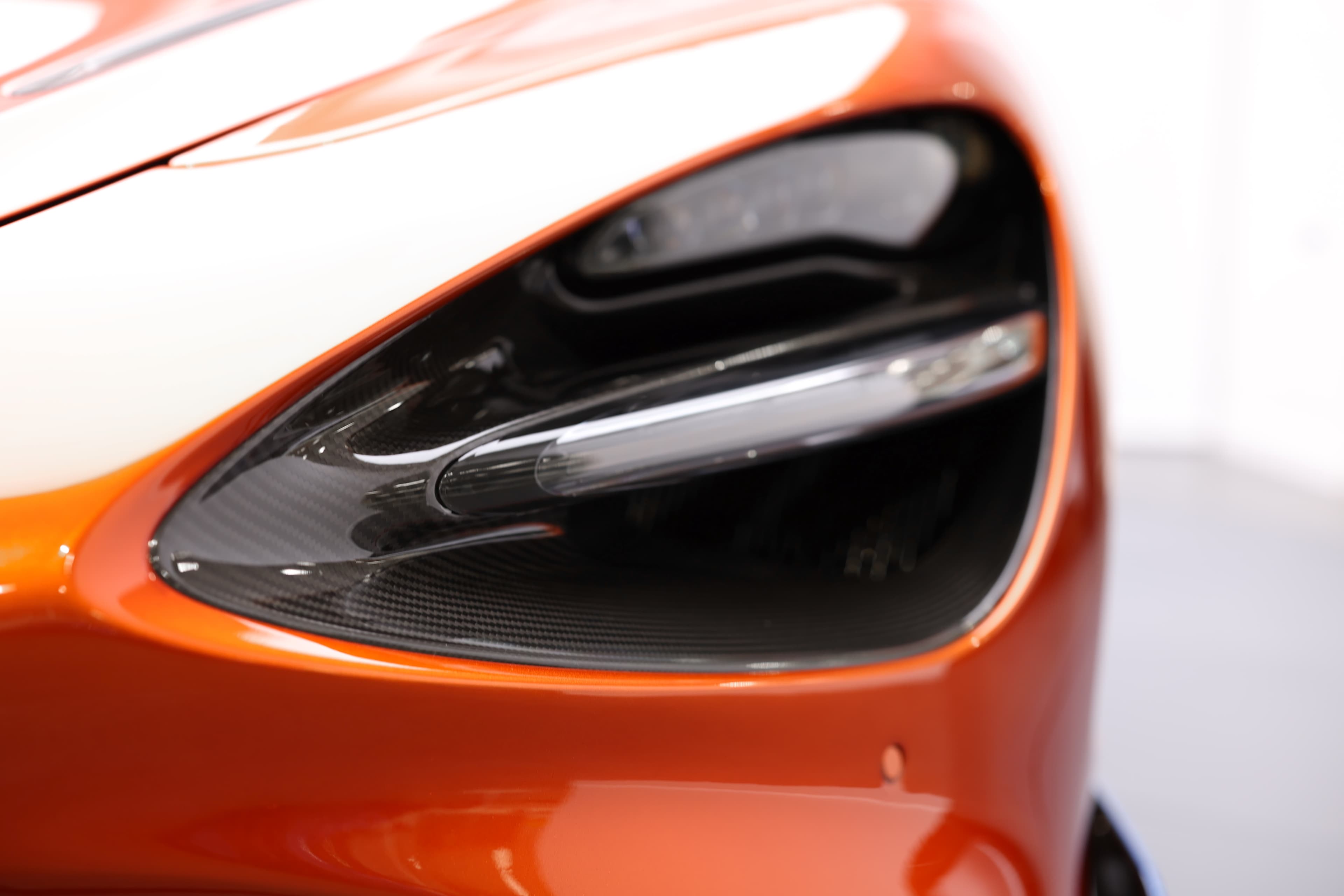 Gros plan sur le phare de la McLaren 765LT, mettant en avant le design dynamique et l'intégration de matériaux en carbone, illustrant l'innovation et l'esthétique moderne de cette supercar.