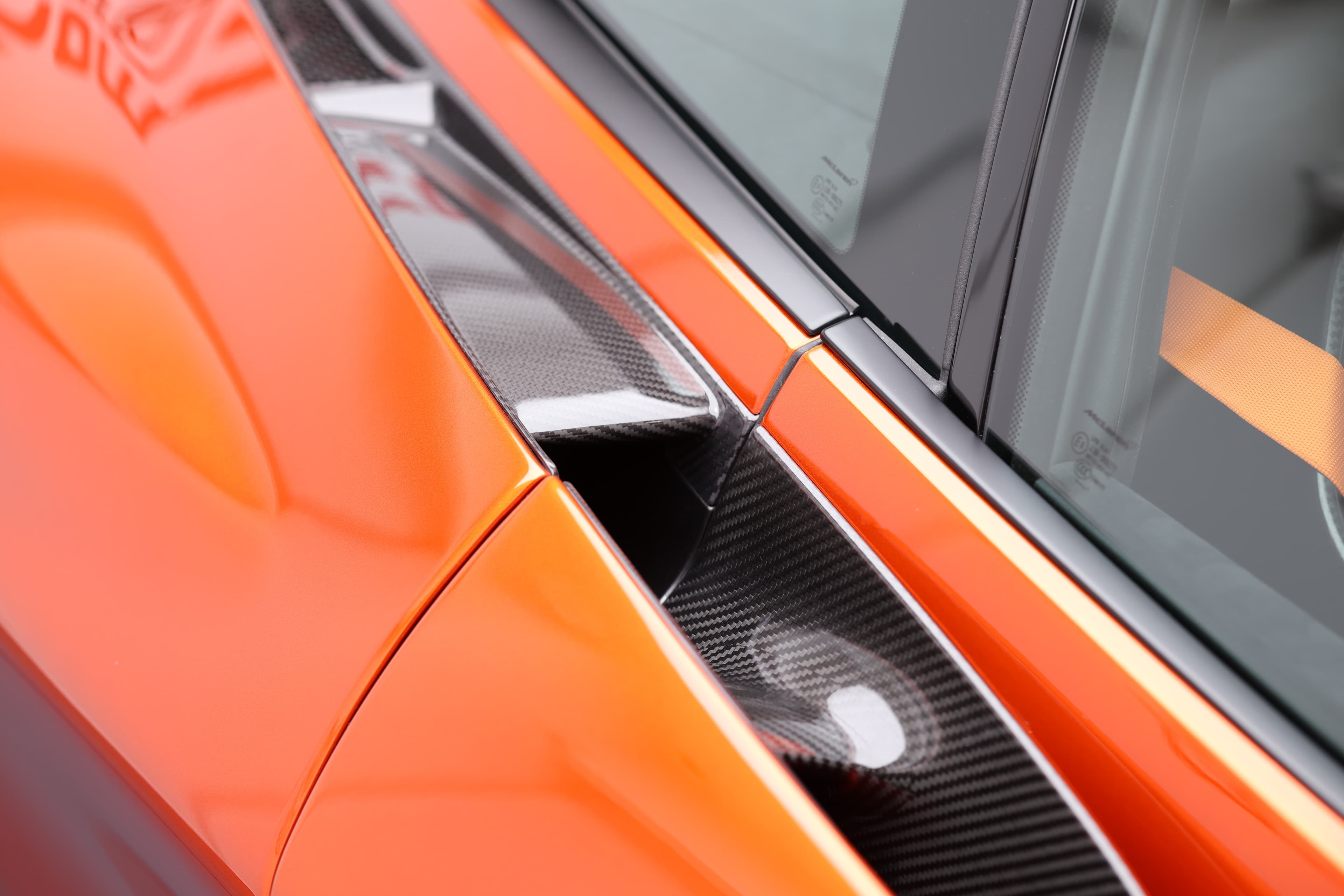 Gros plan sur le détail de la prise d'air latérale de la McLaren 765LT, mettant en avant le contraste entre la peinture orange vive et les éléments en carbone, soulignant l'accent mis sur l'aérodynamisme et le style.
