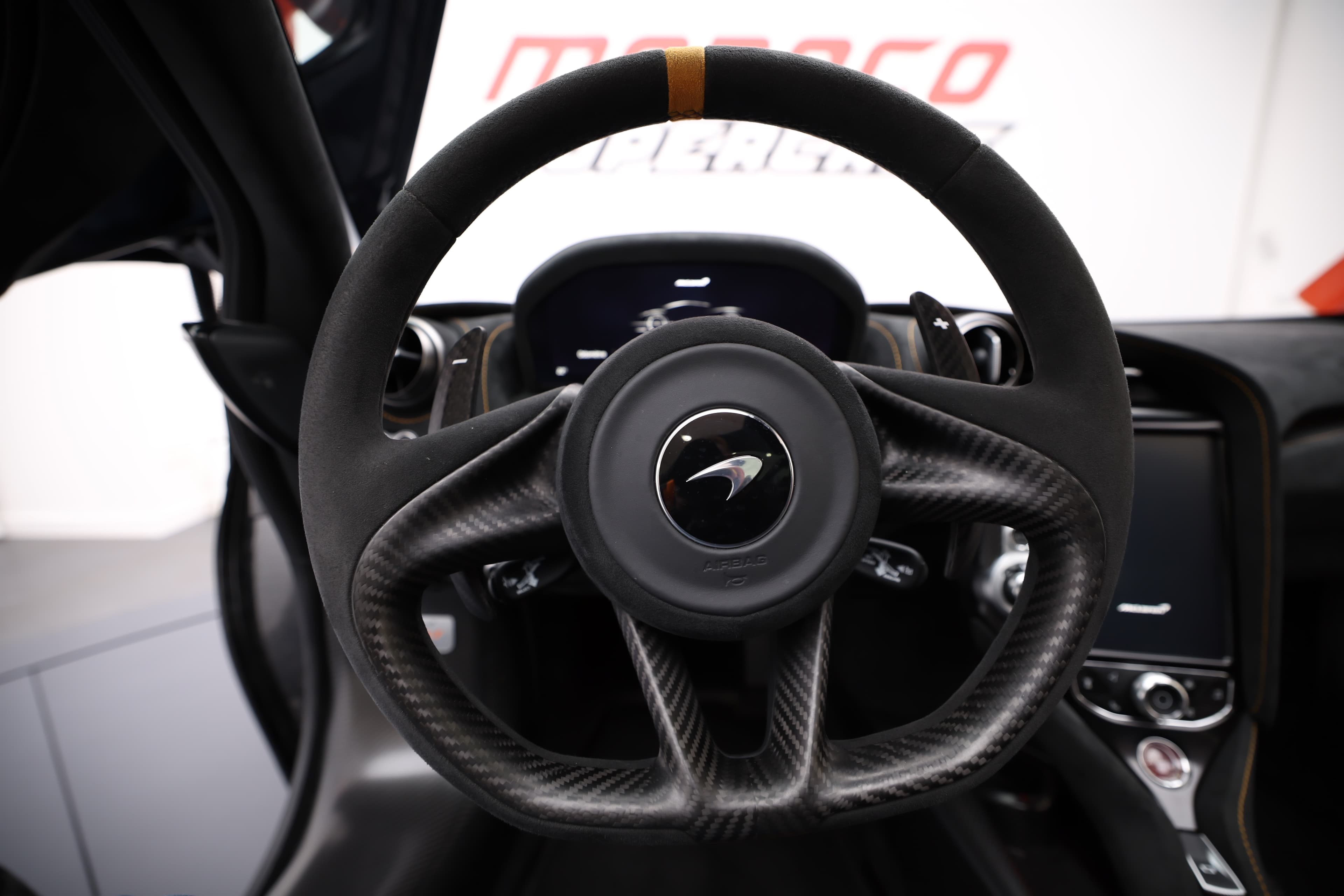 Gros plan sur le volant de la McLaren 765LT, mettant en avant le design ergonomique en carbone et en cuir, ainsi que les commandes intégrées, symbolisant le contrôle et la précision.