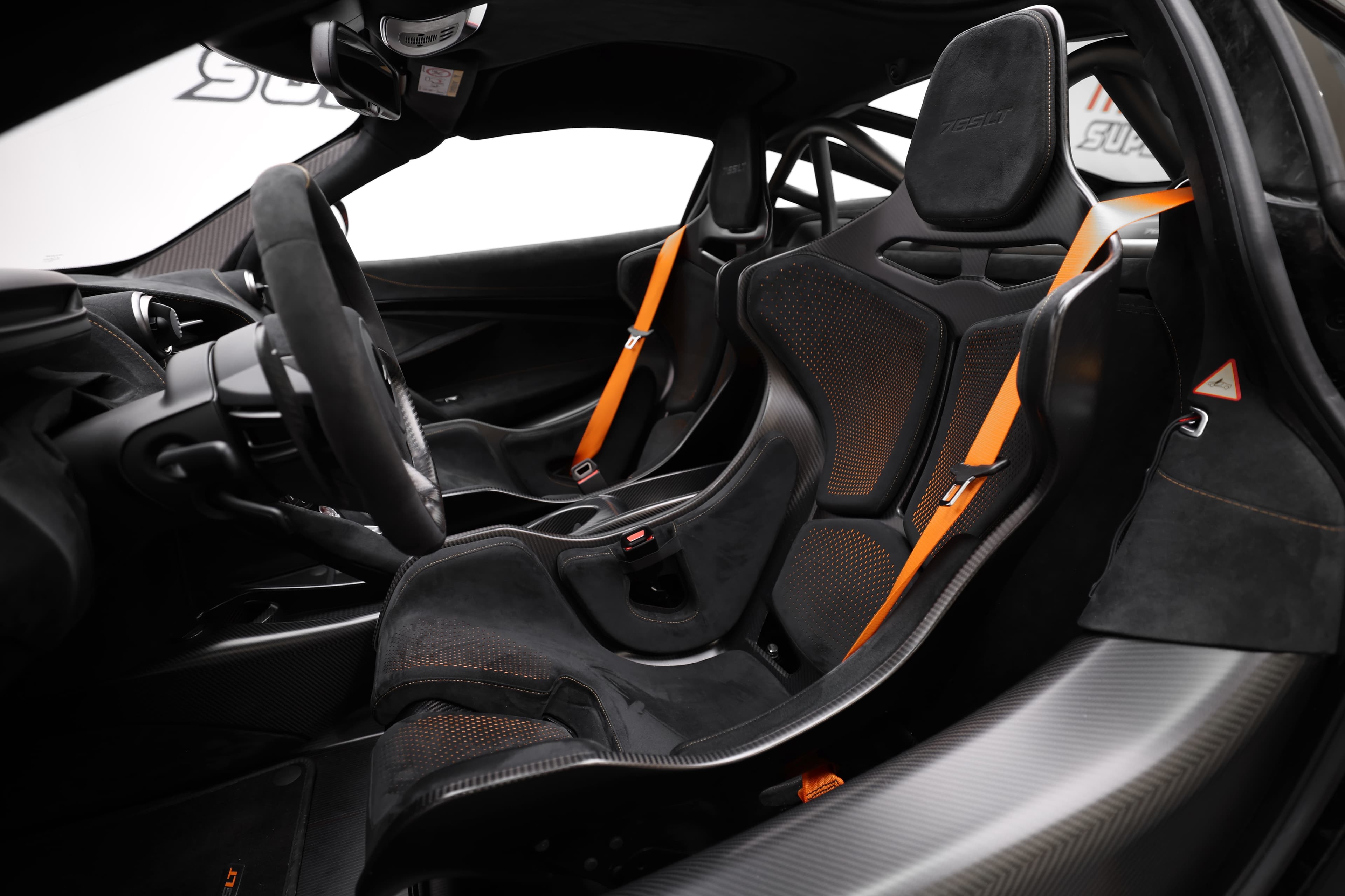 Vue d'ensemble de l'intérieur de la McLaren 765LT, mettant en avant les sièges baquets en carbone, les ceintures de sécurité orange et le design épuré, soulignant l'accent mis sur la performance et le confort.