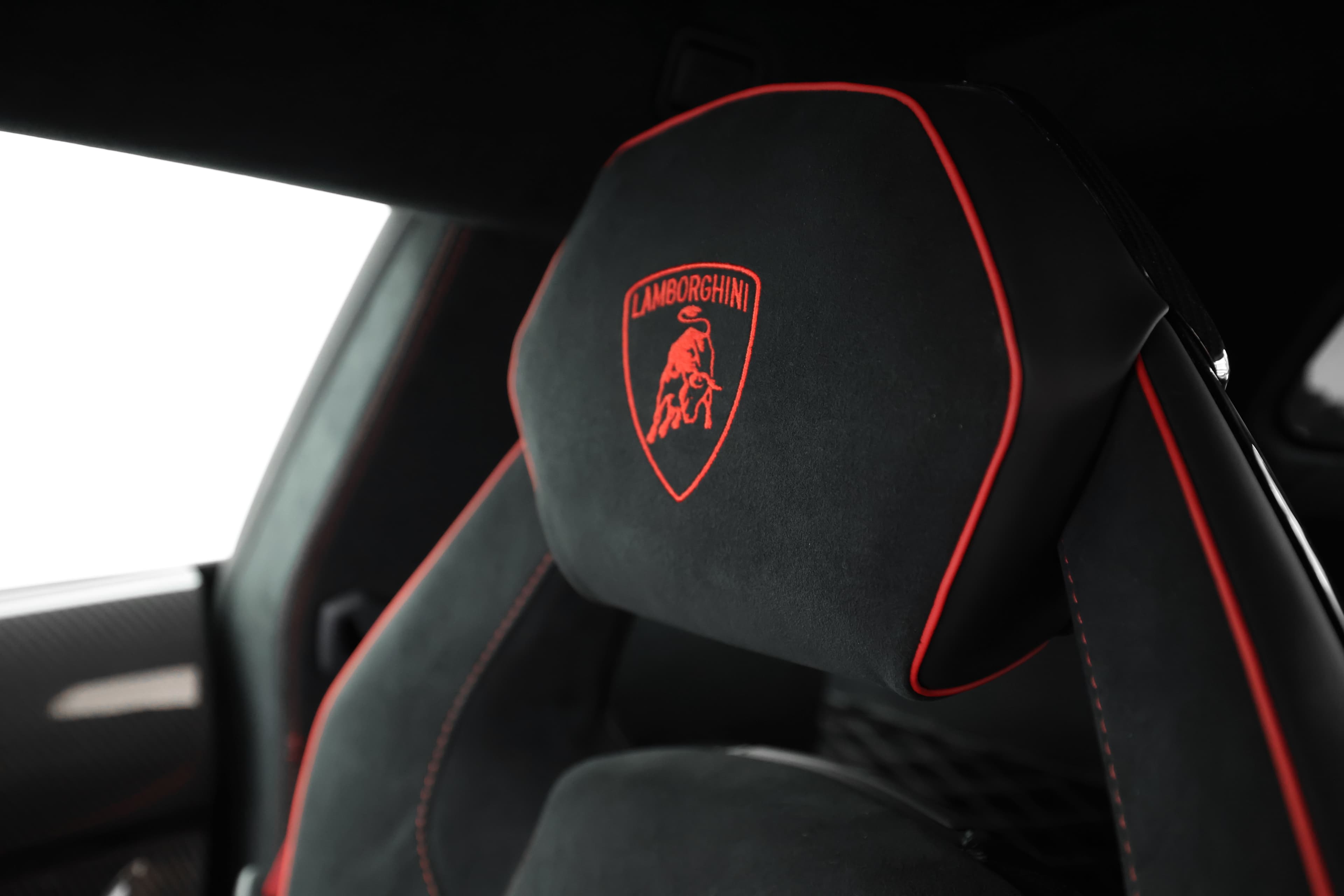 Zoom sur le dossier d'un siège de la Lamborghini Aventador SV, mettant en avant le logo emblématique et les finitions en alcantara. Les lignes rouges ajoutent une touche de sportivité, tandis que le design ergonomique assure un maintien optimal pour une expérience de conduite intense.
