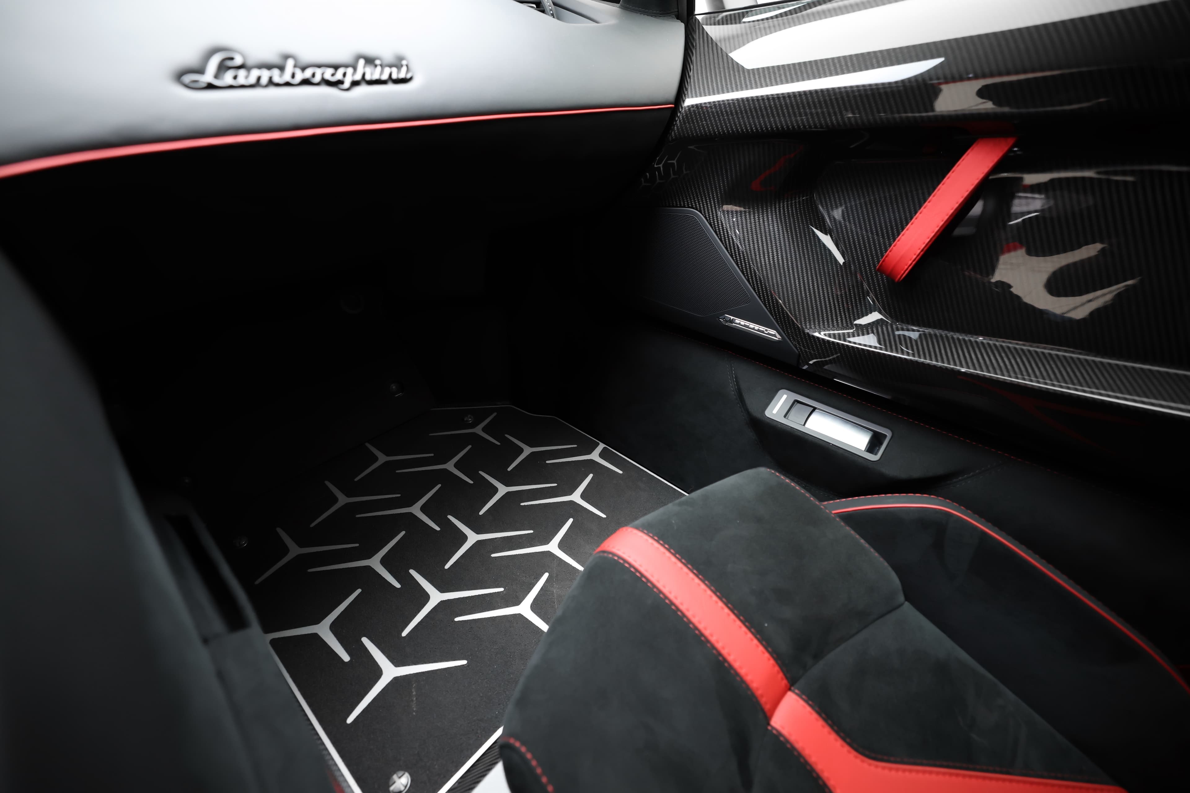 Vue d'ensemble de l'intérieur de la Lamborghini Aventador SV, mettant en avant le logo Lamborghini et les finitions raffinées. Le tapis de sol et les accents rouges ajoutent une touche dynamique, soulignant le mélange de luxe et de sportivité qui caractérise cet emblème de performance.