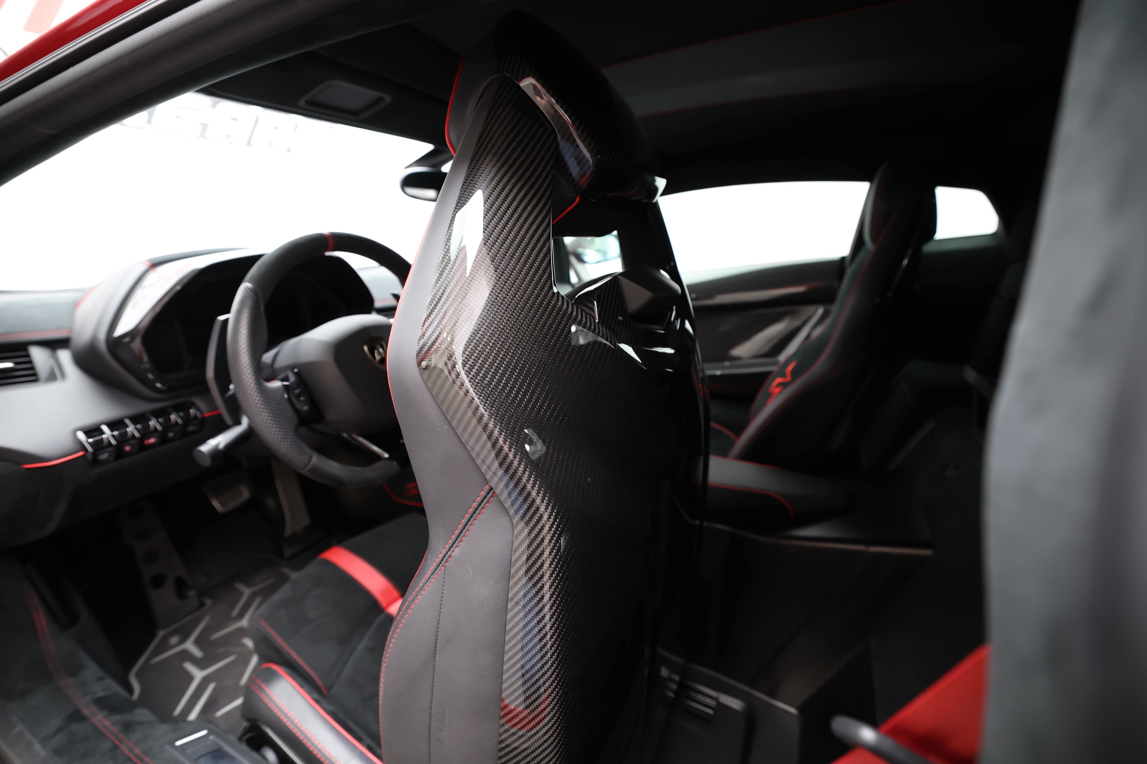 Vue dynamique de l'intérieur de la Lamborghini Aventador SV, axée sur le siège en carbone et le volant. Les éléments en fibre de carbone soulignent la légèreté et la performance, tandis que les détails en rouge créent une ambiance sportive et raffinée, parfaite pour une expérience de conduite exaltante.