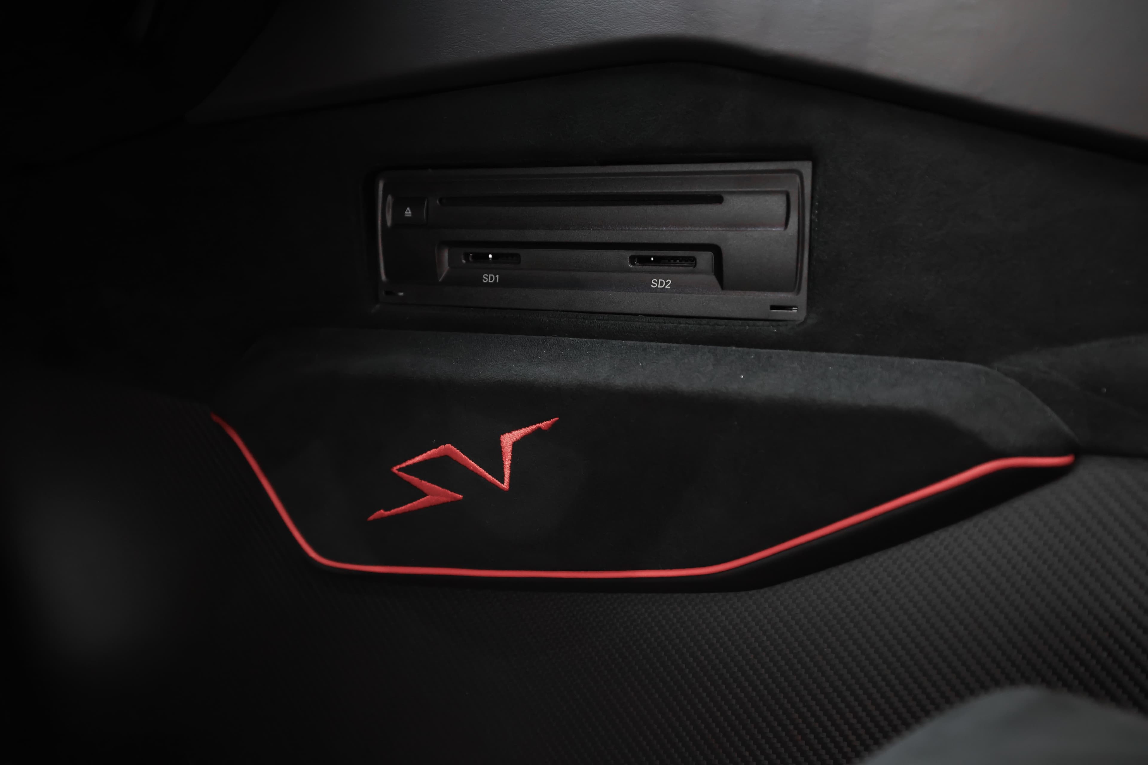 Détail du lecteur de disque intégré dans le panneau de porte de la Lamborghini Aventador SV. Cet élément pratique est entouré de finitions en carbone, avec des accents rouges qui ajoutent une touche de sportivité. Cela démontre comment Lamborghini allie technologie moderne et design raffiné, permettant aux occupants de profiter de leur musique tout en étant immergés dans un environnement luxueux